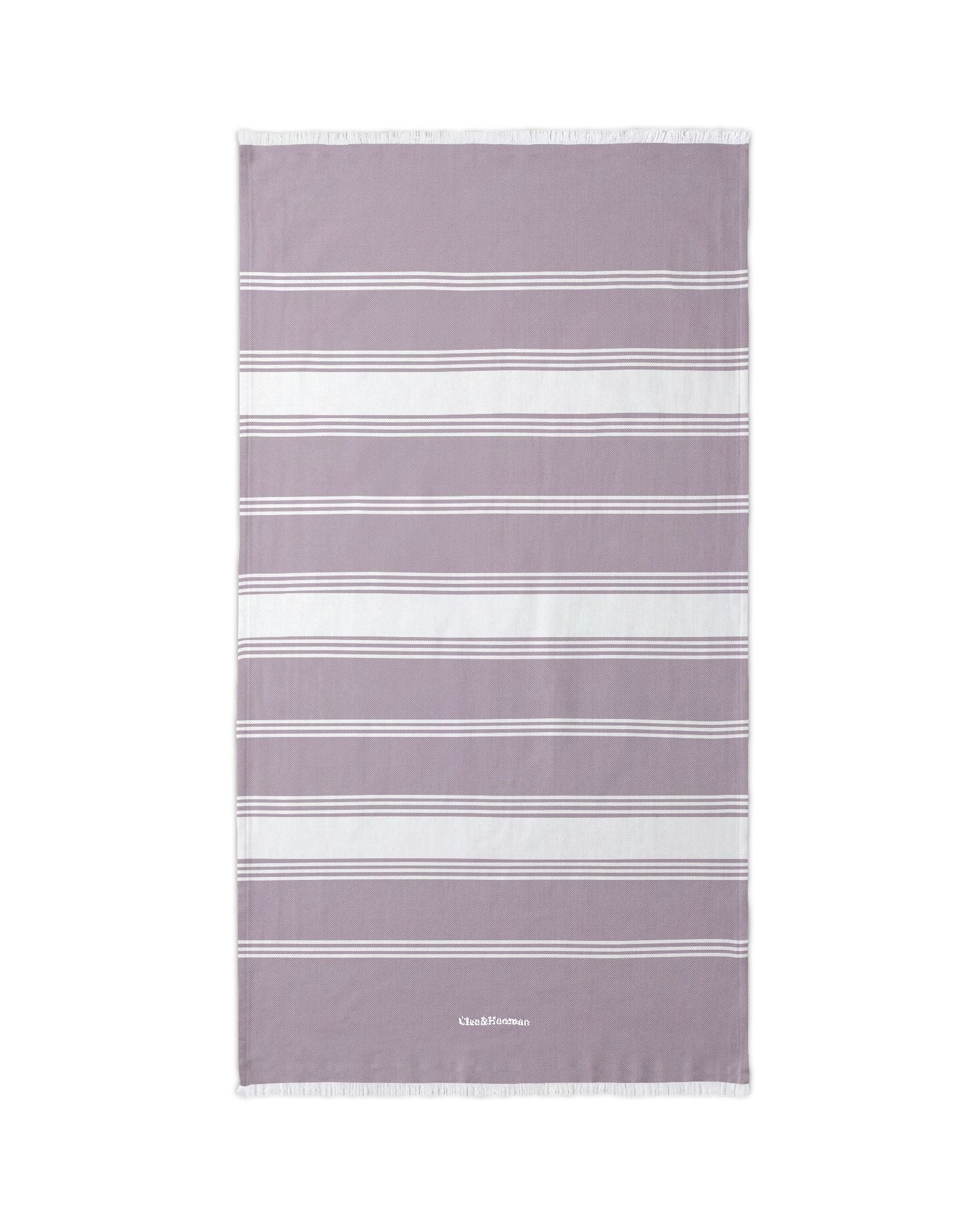 A rectangular beach towel with horizontal white and light gray stripes on a dusty lavender-purple background, featuring a small white embroidered logo at the bottom and white fringe on the top and bottom edges.