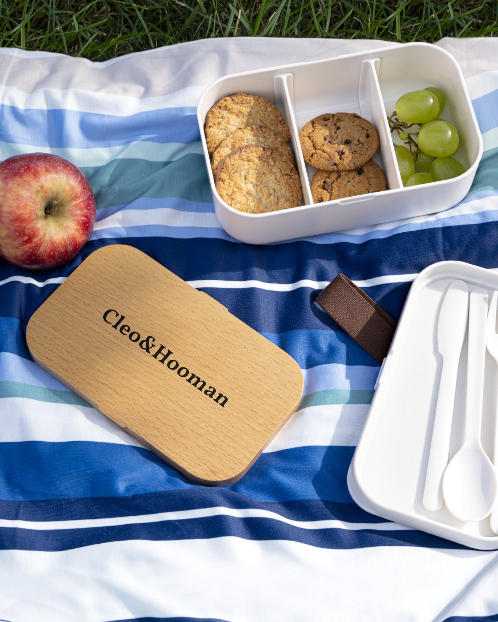 Picnic setup with a wooden board, food containers, and utensils on a striped blanket outdoors.