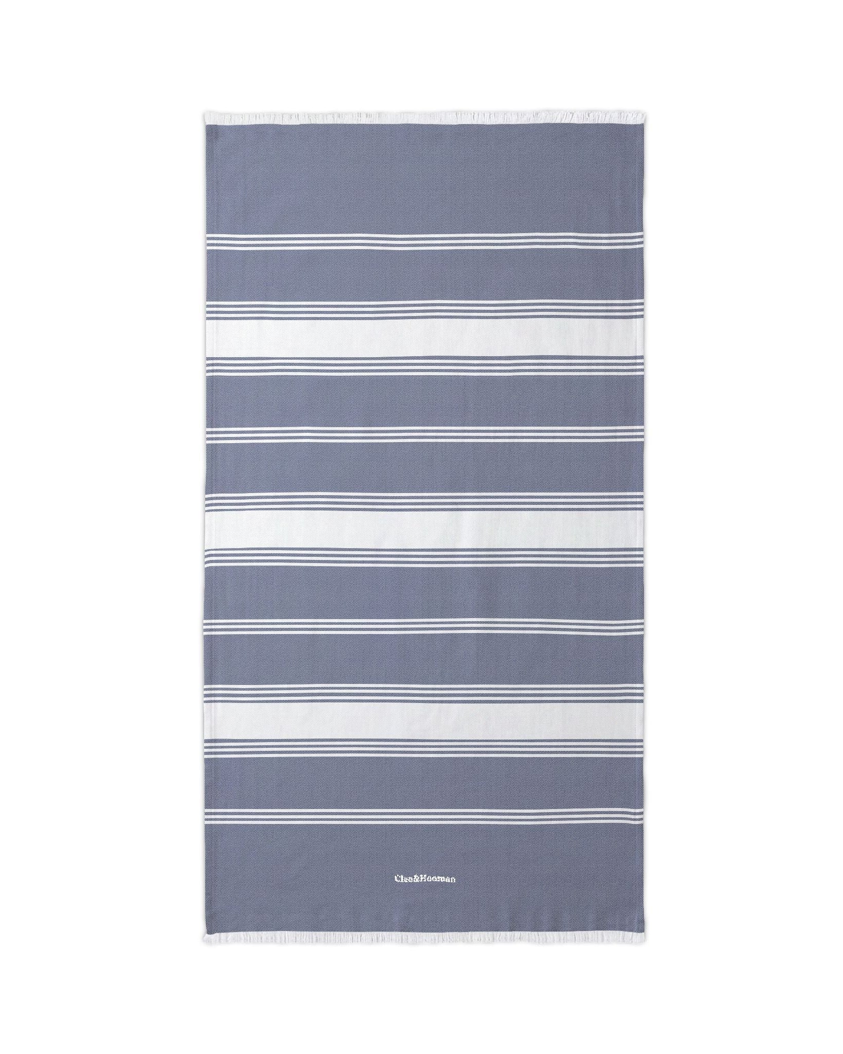 A rectangular beach towel with horizontal stripes in slate blue, white, and light gray, featuring a small white 'TheBeachGoods' logo near the bottom edge.