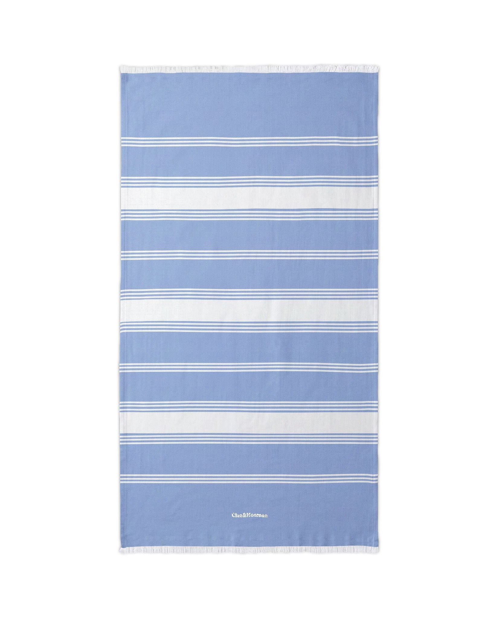A rectangular beach towel with horizontal stripes in light blue, white, and navy, featuring a small white logo at the bottom center, displayed flat against a solid white background.