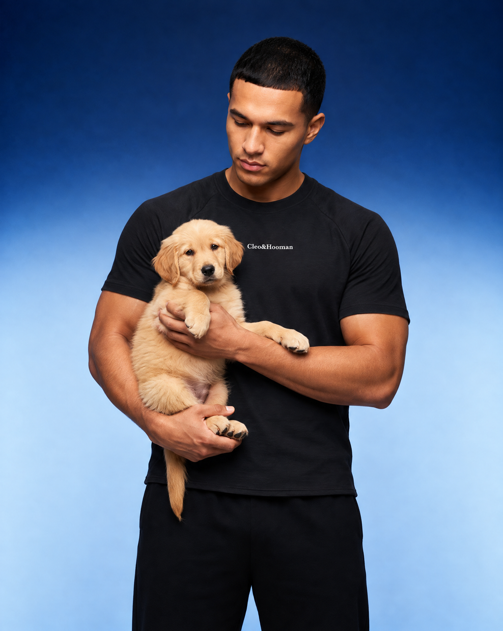 A man wearing a black t-shirt and black pants holds a golden retriever puppy against a blue gradient studio background. The t-shirt features the text 'Cleo&Hooman' printed in white on the left chest.