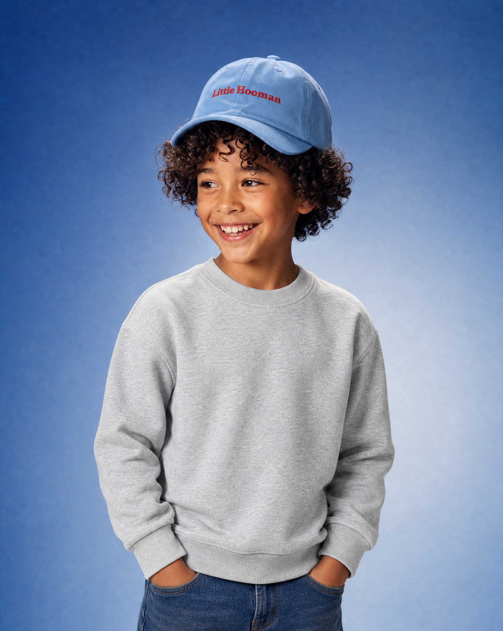 A young boy with curly hair smiling, wearing a light gray crewneck sweatshirt, blue jeans, and a light blue baseball cap with 'Little Hooman' embroidered in red on the front, set against a blue gradient studio background.