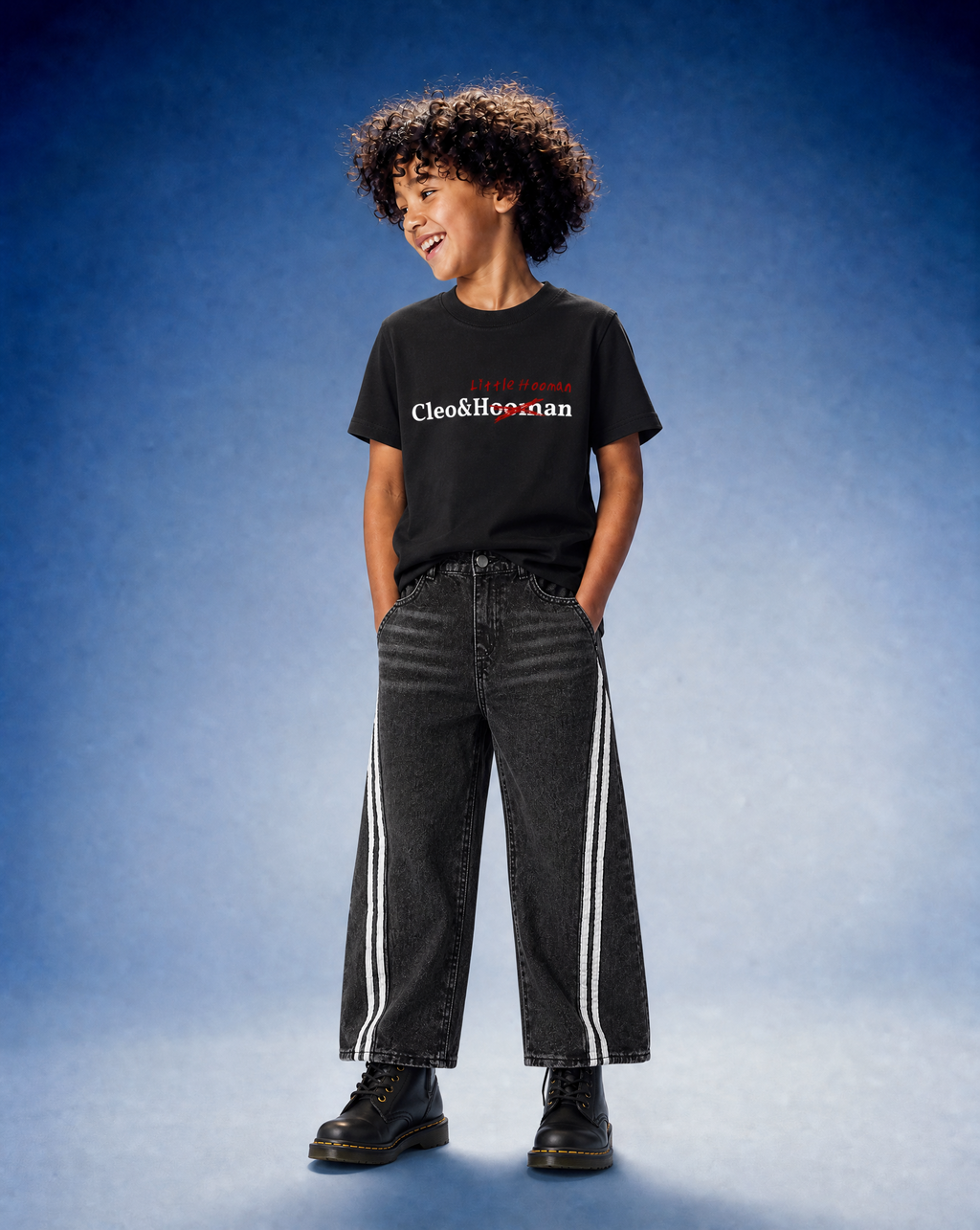 A child model wearing a black Cleo & Honzan graphic t-shirt, dark gray wide-leg jeans with white side stripes, and black boots, standing against a blue studio backdrop.