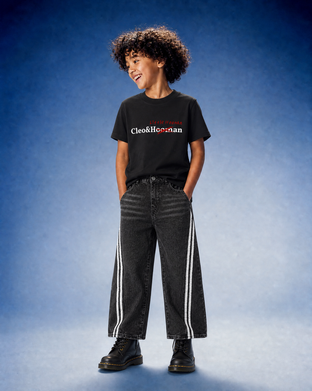 A child model wearing a black Cleo & Honzan graphic t-shirt, dark gray wide-leg jeans with white side stripes, and black boots, standing against a blue studio backdrop.