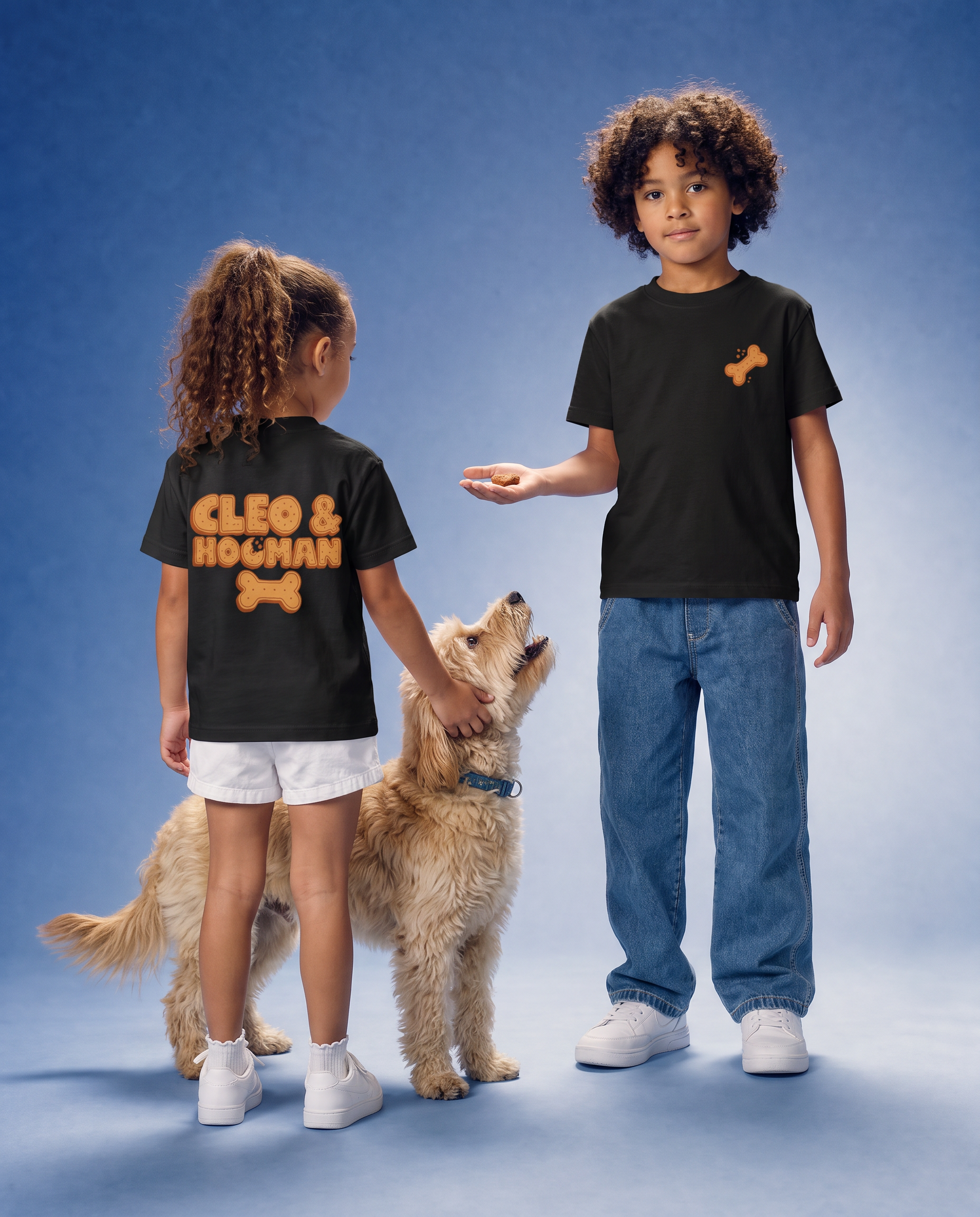 Two children modeling black graphic t-shirts with a dog design, standing next to a golden dog against a blue studio backdrop. The child on the left shows the back of the shirt with 'CLEO & Hooman' and a bone graphic, while the child on the right shows the front chest with a small bone graphic.
