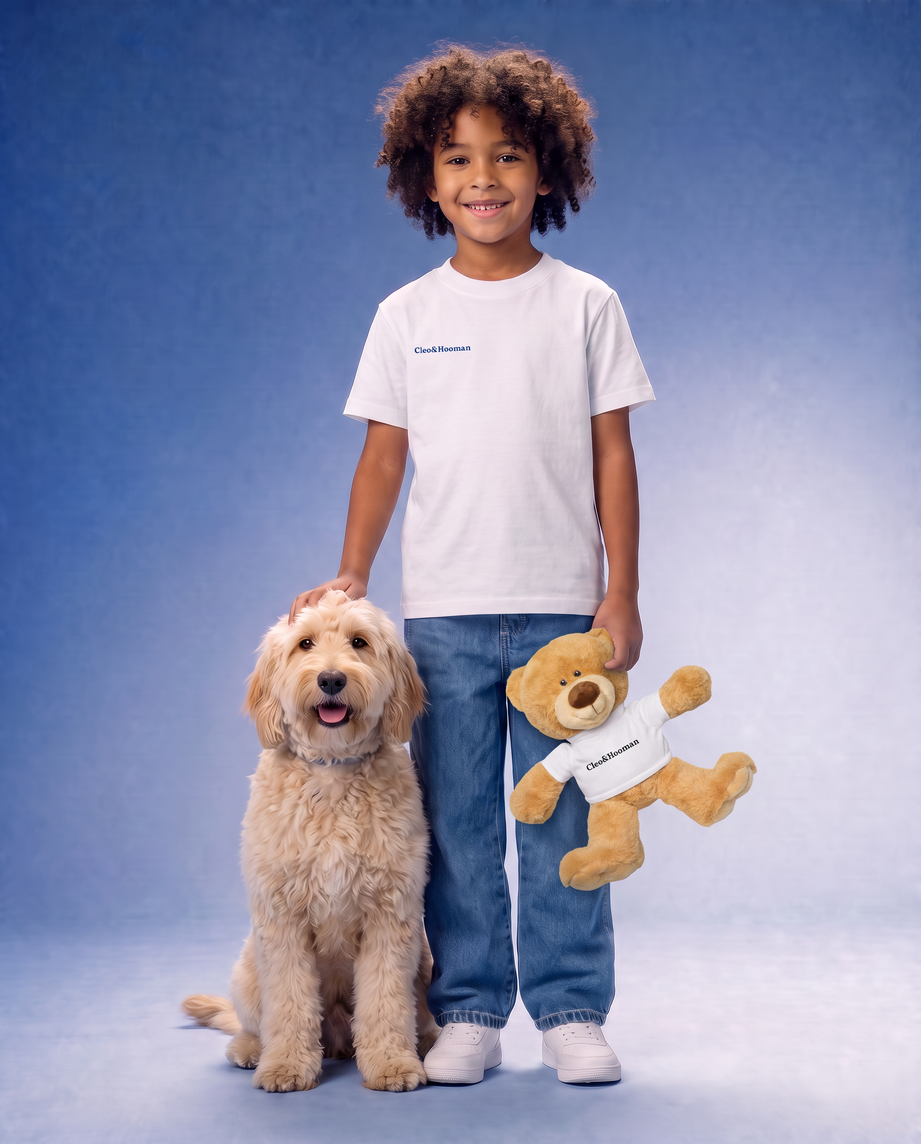 A young boy with curly hair stands next to a fluffy cream-colored dog, wearing a white t-shirt with a small blue 'Cleo&Hooman' logo. He is holding a plush brown teddy bear that also has a white 'Cleo&Hooman' t-shirt.