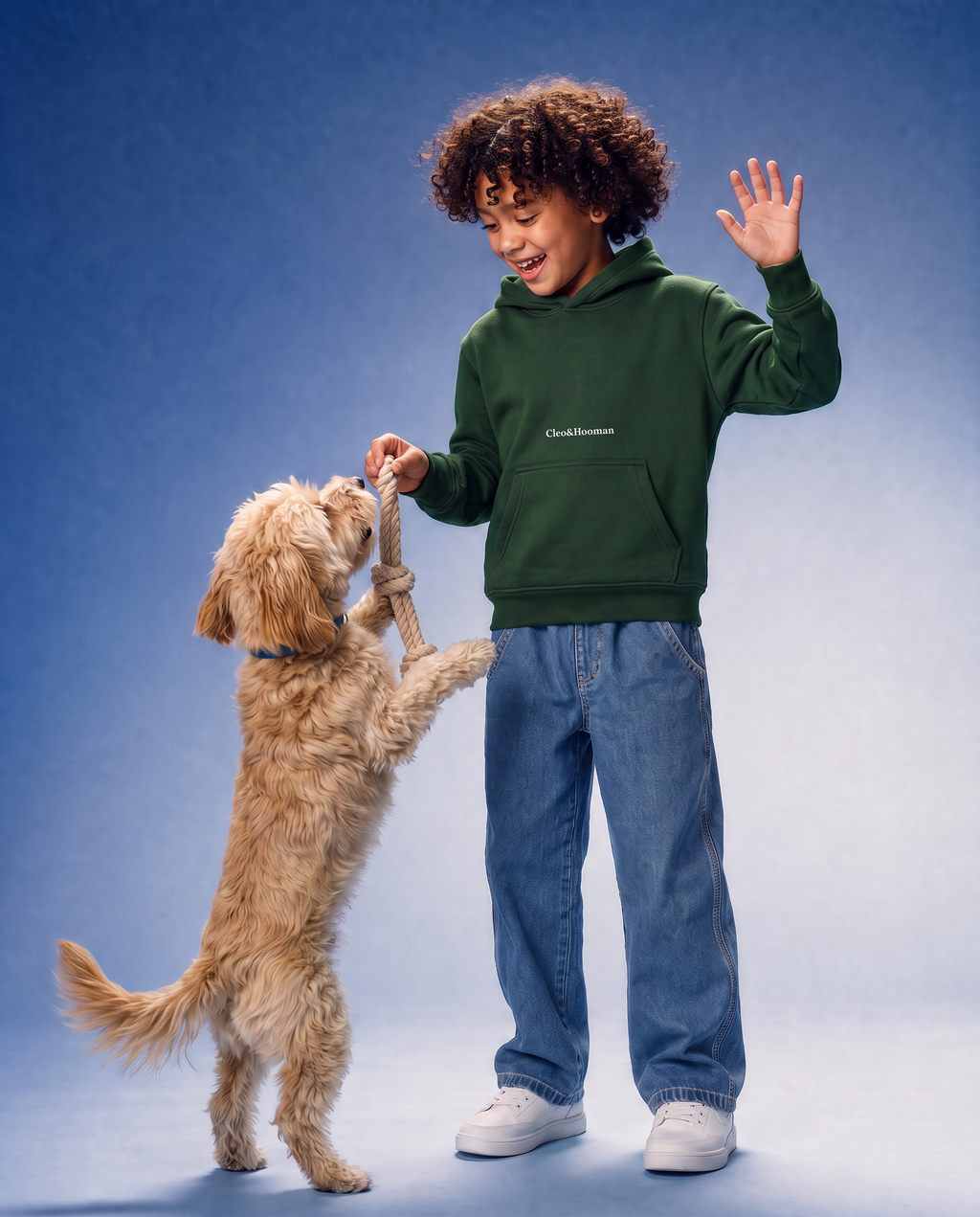A child wearing a dark green hoodie with 'cleo&hooman' printed on the chest, blue jeans, and white sneakers is playing tug-of-war with a small golden dog on a blue studio background.