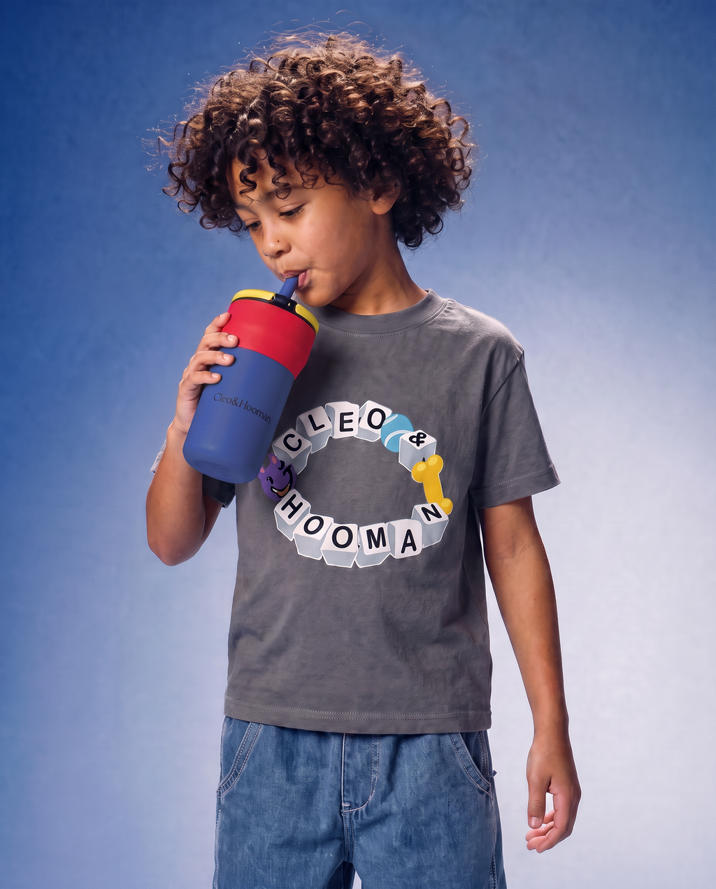 A child with curly brown hair wearing a gray graphic t-shirt and blue jeans, drinking from a blue and red water bottle against a blue gradient background.