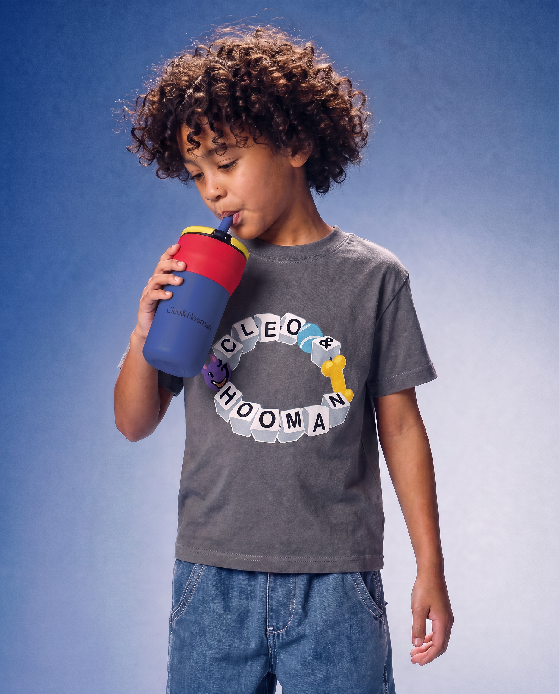A child with curly brown hair wearing a gray graphic t-shirt and blue jeans, drinking from a blue and red water bottle against a blue gradient background.