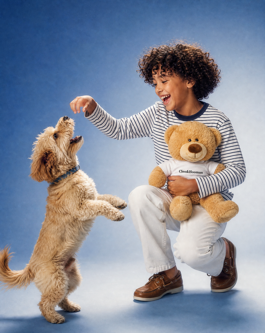 A smiling child crouches on a blue studio background, reaching toward a small fluffy dog while holding a teddy bear. The child wears a striped long-sleeve shirt, white pants, and brown shoes, and the teddy bear wears a white shirt with the visible brand name 'Cleo&Hooman'.