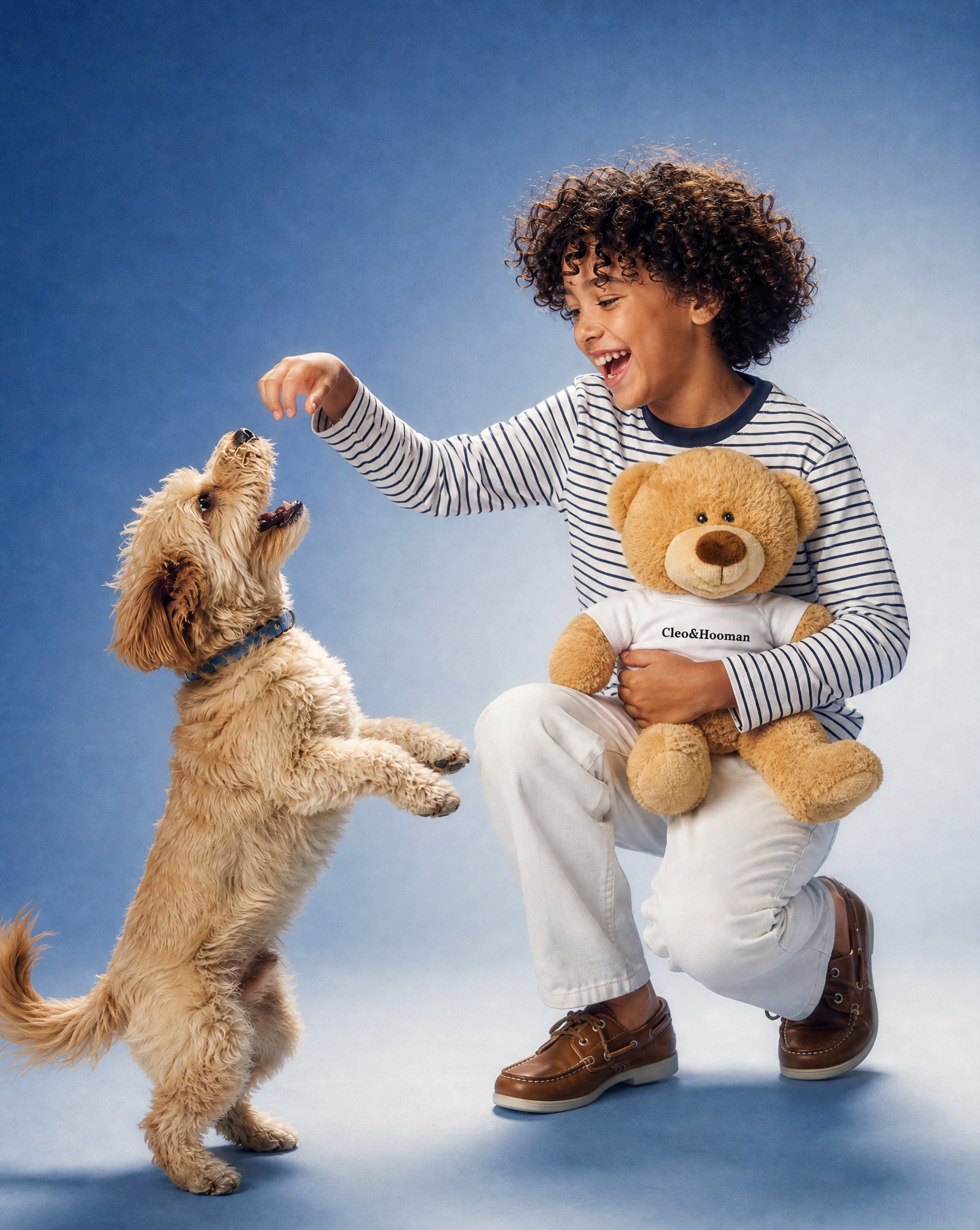 A smiling child crouches on a blue studio background, reaching toward a small fluffy dog while holding a teddy bear. The child wears a striped long-sleeve shirt, white pants, and brown shoes, and the teddy bear wears a white shirt with the visible brand name 'Cleo&Hooman'.