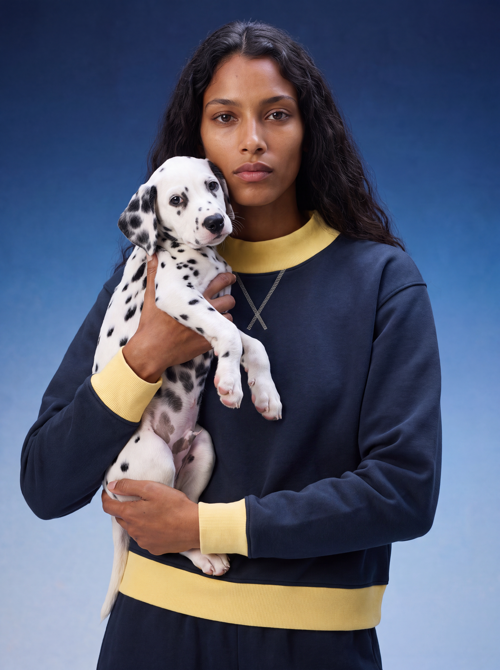A woman holding a Dalmatian puppy, wearing a navy blue sweatshirt with yellow ribbed trim and a matching navy and yellow sweatpants set, against a blue studio background.