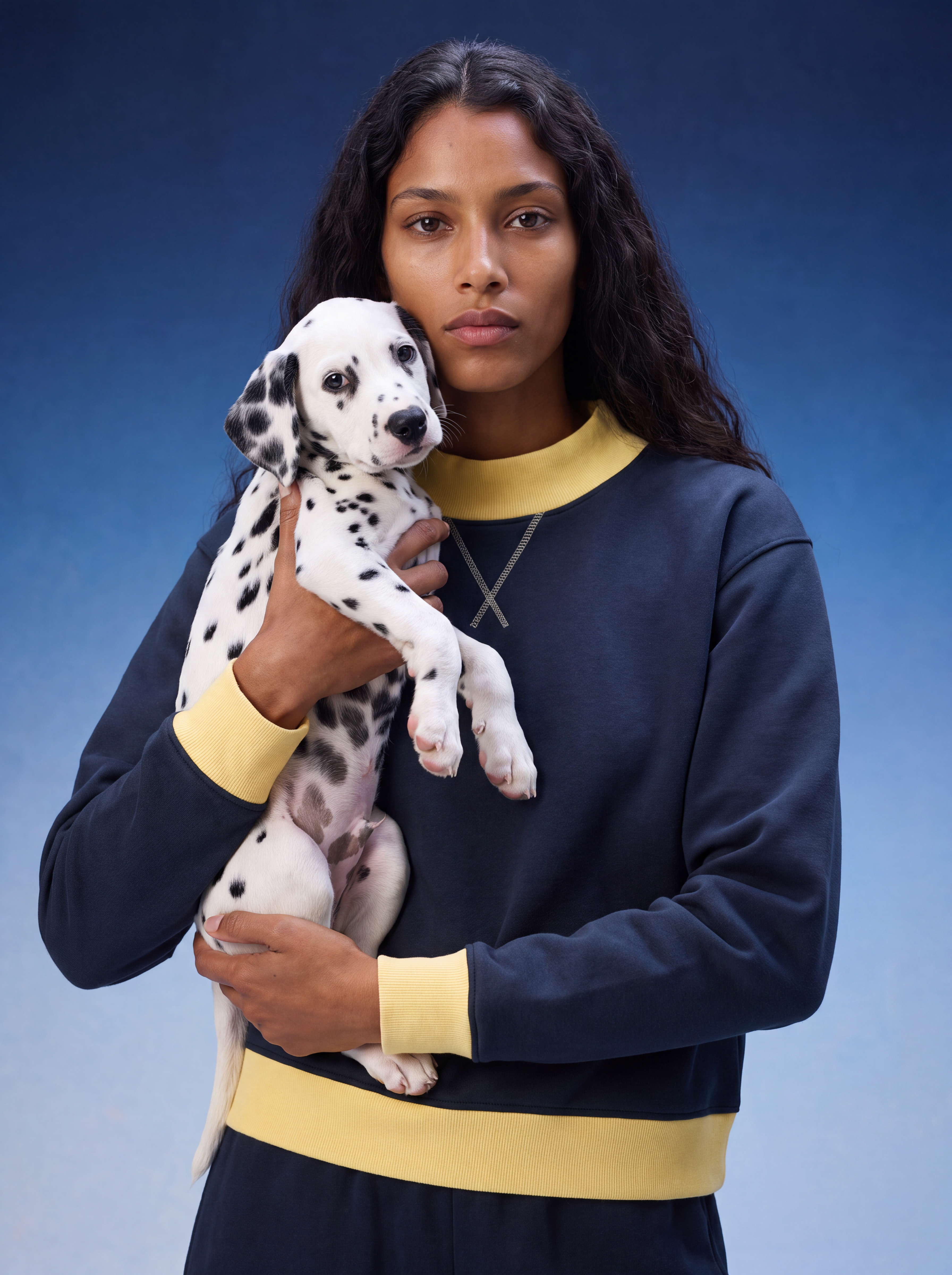 A woman holding a Dalmatian puppy, wearing a navy blue sweatshirt with yellow ribbed trim and a matching navy and yellow sweatpants set, against a blue studio background.
