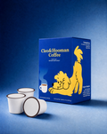 A blue box of Cleo & Hooman Coffee with a yellow cartoon dog illustration stands on a blue studio background, with three white coffee pods placed in front.