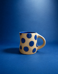 A handmade ceramic mug with a wavy silhouette, featuring a beige speckled exterior decorated with large blue polka dots, and a solid blue interior, set against a solid blue background.