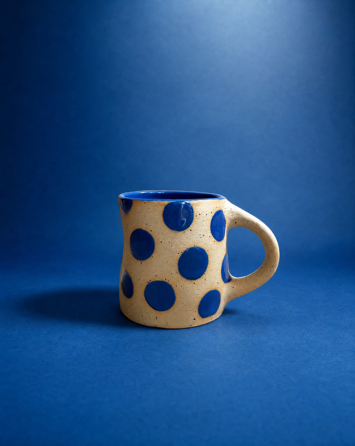 A handmade ceramic mug with a wavy silhouette, featuring a beige speckled exterior decorated with large blue polka dots, and a solid blue interior, set against a solid blue background.